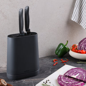 The Zyliss Universal Knife Block, in black with a flexible bristle insert and two knives, sits on a kitchen counter next to purple cabbage, green bell pepper, red chili peppers, and a striped towel.