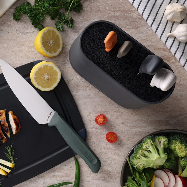On the kitchen counter, a Zyliss 6 Piece Universal Knife Block with stainless steel knives and ergonomic handles sits beside sliced lemon, grilled chicken, cherry tomatoes, broccoli, herbs, a striped towel, and garlic cloves.
