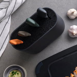 The Zyliss 6 Piece Universal Knife Block, featuring three stainless steel knives with ergonomic orange, gray, and dark green handles, sits on a gray countertop beside garlic bulbs, a striped towel, and mashed potatoes.
