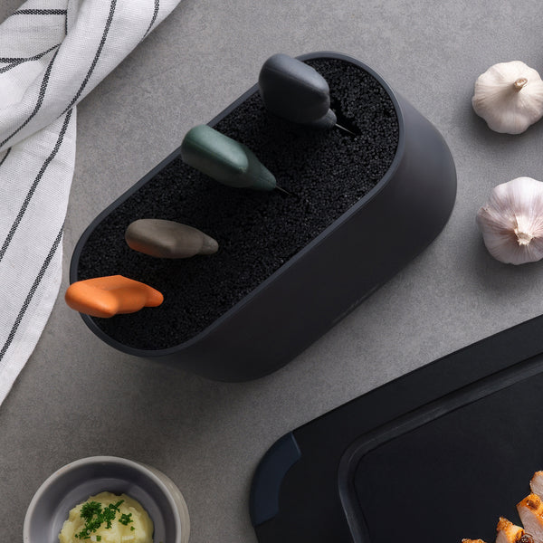 The Zyliss 6 Piece Universal Knife Block, featuring three stainless steel knives with ergonomic orange, gray, and dark green handles, sits on a gray countertop beside garlic bulbs, a striped towel, and mashed potatoes.