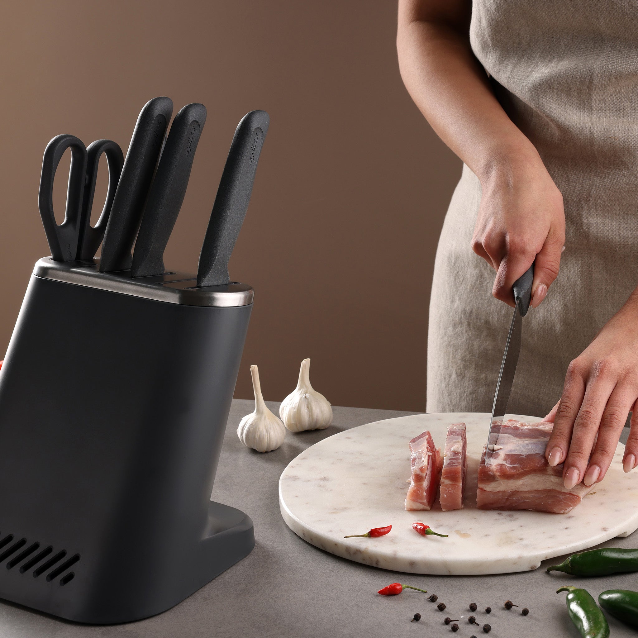 Someone slices raw meat on a round marble cutting board beside a Zyliss 8-Piece Knife Block holding knives and scissors, with garlic bulbs, chili peppers, and peppercorns scattered nearby.