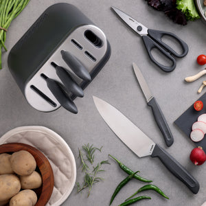The Zyliss 8-Piece Knife Block includes sustainable knives, kitchen scissors, and a cutting board with chopped veggies, green chilies, fresh herbs, potatoes in a bowl, plus a potholder—all on a gray countertop.