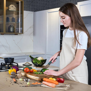 A woman in a beige apron uses the Zyliss 3 Piece Knife Set to slice green bell pepper on a wooden cutting board in a modern kitchen, surrounded by fresh vegetables such as carrots, tomatoes, and mushrooms.