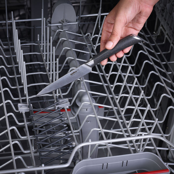 A hand places a Zyliss Comfort Pro Paring Knife with a dark handle from the Comfort Pro Paring Knife Set into the top rack of an open dishwasher, ready for the next round of kitchen prep.
