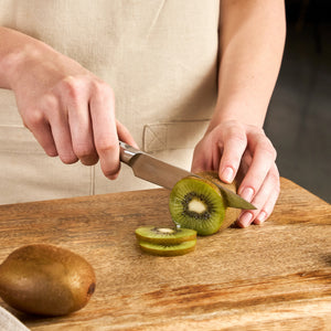 A person slices a kiwi with the Zyliss Comfort Pro Paring Knife 11cm / 4½" on a wooden cutting board while wearing a beige apron. One whole kiwi and two kiwi slices are visible.