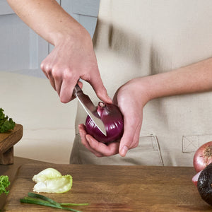 A person stands by a wooden table with lettuce and other vegetables, holding a red onion and slicing it with the Zyliss Comfort Pro Paring Knife 11cm / 4½".