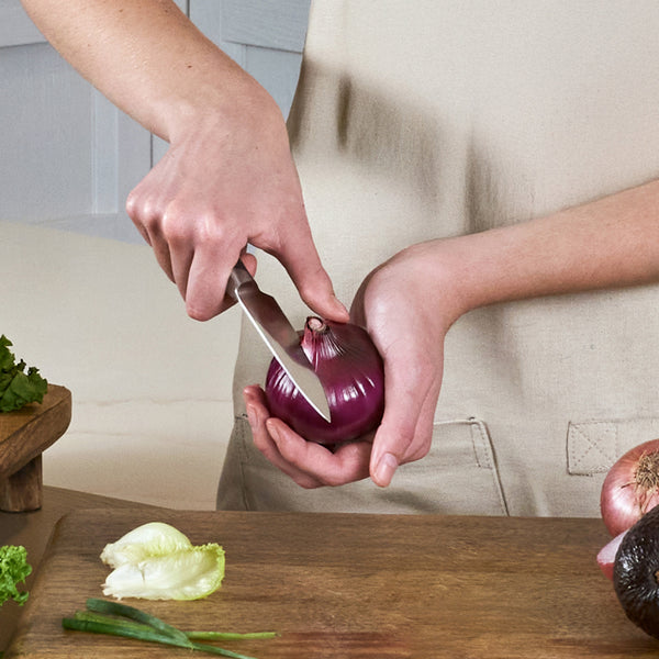 A person stands by a wooden table with lettuce and other vegetables, holding a red onion and slicing it with the Zyliss Comfort Pro Paring Knife 11cm / 4½".
