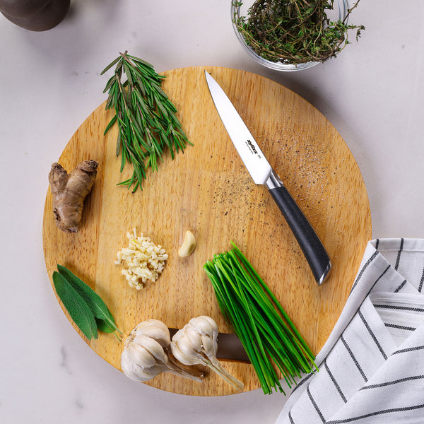 A round wooden cutting board with the Zyliss Comfort Pro Paring Knife 11cm/4½", fresh rosemary, thyme, ginger, sage, chopped and whole garlic, chives, and a striped kitchen towel on a light countertop.