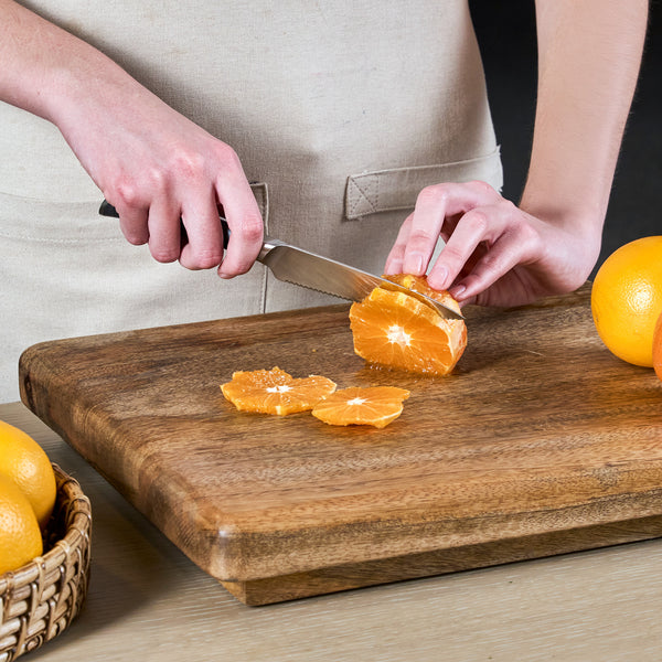 A person slices an orange with the Zyliss Comfort Pro Serrated Paring Knife 11.5cm, which features an ergonomic handle, on a wooden cutting board while a basket of whole oranges sits nearby on the table.