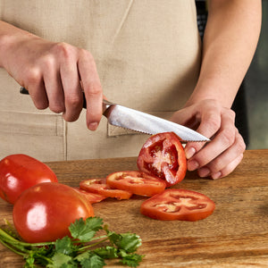 Wearing a beige apron, a person uses the Zyliss Comfort Pro Paring Knife Set to slice a tomato on a wooden cutting board during kitchen prep. Tomato slices, whole tomatoes, and fresh cilantro are arranged on the board.