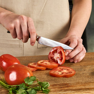 Wearing a beige apron, a person slices a tomato on a wooden board using the Zyliss Comfort Pro Serrated Paring Knife 11.5cm / 4½" with an ergonomic handle; sliced and whole tomatoes plus cilantro are also on the board.