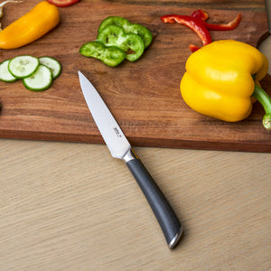 The Zyliss Comfort Pro Serrated Paring Knife 11.5cm with an ergonomic black handle rests on a wooden surface beside a cutting board topped with sliced cucumber, green and red bell peppers, a yellow bell pepper, and a yellow chili pepper.