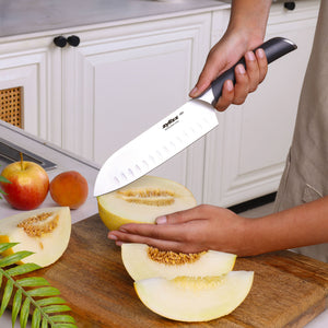 A person slices a yellow melon on a wooden cutting board using the Zyliss Comfort Pro Santoku Knife 18.5cm / 7". Apples and part of a plant rest nearby, with white kitchen cabinets in the background.