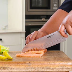 A person slices salmon on a wooden cutting board using the Zyliss Comfort Pro Santoku Knife 18.5cm/7" with an ergonomic handle. Slices of lemon and lettuce are nearby, set in a modern kitchen backdrop.