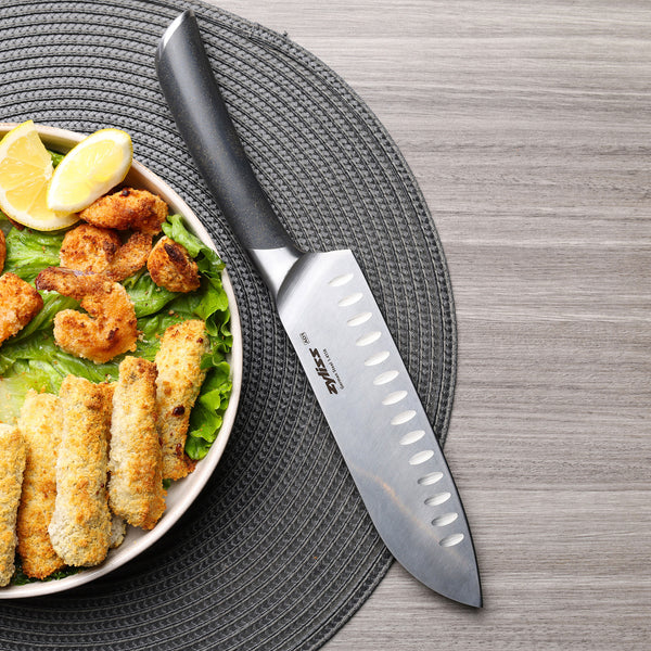 The Zyliss Comfort Pro Santoku Knife 18.5cm / 7" with a black ergonomic handle rests on a gray woven placemat next to baked food, green lettuce, and lemon wedges, arranged on a light gray wooden table.