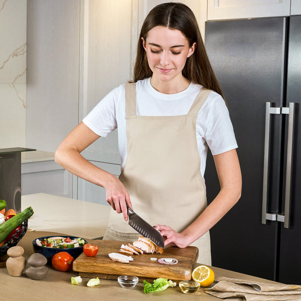 A woman in a beige apron slices cooked chicken with the Zyliss Comfort Pro Santoku Knife 18.5cm/7" on a wooden board in a modern kitchen, surrounded by fresh vegetables, a salad bowl, and lemon on the counter.