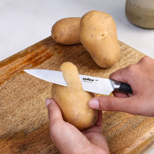 Someone uses a Zyliss Comfort Pro Utility Knife 11.5cm/4½" to peel a potato on a wooden cutting board, with two unpeeled potatoes and a jar in the background.