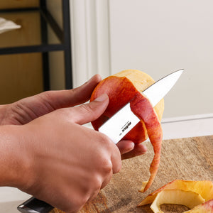 A person uses a Zyliss Comfort Pro Prep Trio Knife Set paring knife to peel the skin off a red apple over a wooden surface, with a long curl of apple peel hanging from the fruit.