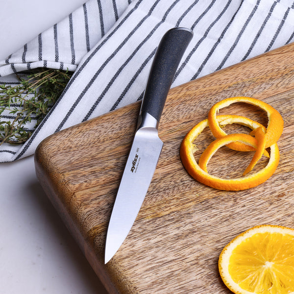 The Zyliss Comfort Pro Utility Knife 11.5cm with a black handle sits on a wooden cutting board next to orange peels, an orange slice, and a striped kitchen towel holding fresh herbs.