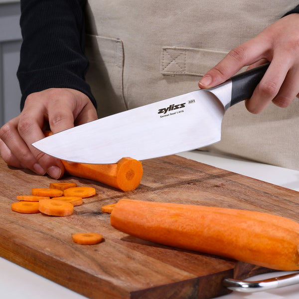Wearing a beige apron and black long-sleeve shirt, a person slices a carrot on a wooden cutting board using the Zyliss Comfort Pro Chef Knife 19cm / 7½". Sliced carrot pieces are scattered on the board.