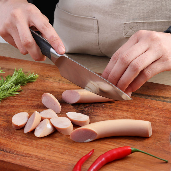 A person uses the Zyliss Comfort Pro Chef + Santoku Knife Set to slice sausages on a wooden cutting board, with rosemary and two red chili peppers also arranged on the board.