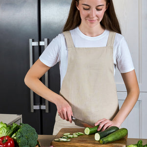 A woman in a beige apron slices a cucumber with the Zyliss Comfort Pro Chef Knife 19cm / 7½", using its ergonomic handle on a wooden board, surrounded by fresh vegetables like broccoli, lettuce, and red pepper in her kitchen.