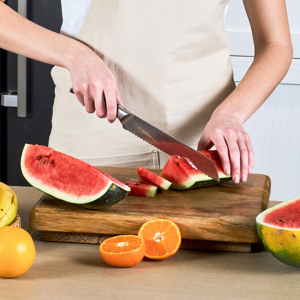 Wearing a beige apron, a person slices a watermelon on a wooden cutting board using the Zyliss Comfort Pro Bread Knife 21.5cm / 8½" with its serrated blade. Orange slices and assorted fruits are nearby.