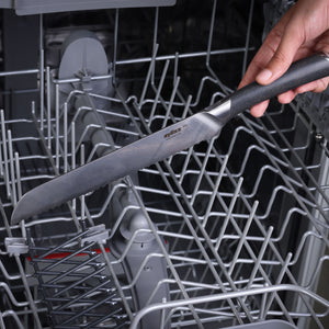 A hand places a large knife from the Zyliss Comfort Pro Kitchen Quartet Knife Set, featuring a black handle, into an empty dishwasher rack.