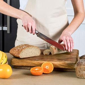 Wearing a beige apron, a person slices whole grain bread on a wooden cutting board with the Zyliss Comfort Pro Bread Knife 21.5cm/8½", surrounded by oranges, a banana, and extra bread on the kitchen counter.