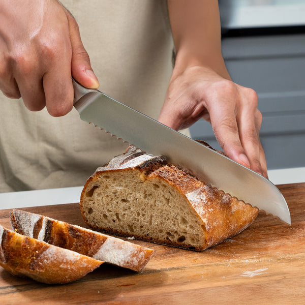 Using a serrated knife from the Zyliss Comfort Pro Kitchen Quartet Knife Set, a person slices a loaf of rustic, crusty bread on a wooden cutting board. Two slices rest beside the partially sliced loaf.