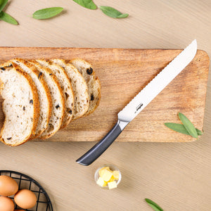 A sliced loaf of raisin bread rests on a wooden board with the Zyliss Comfort Pro Bread Knife 21.5cm beside it. Nearby are a bowl of butter cubes, fresh sage leaves, and a basket of eggs on a light wood surface.