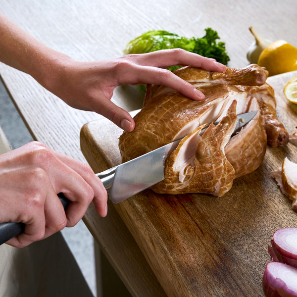 Using the Zyliss Comfort Pro Carving Knife 20cm/8", a person slices smoked chicken breast on a wooden cutting board, surrounded by fresh veggies such as lettuce, red onion, and lemon.