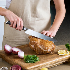 A person in a beige apron slices a roast on a wooden cutting board using the Zyliss Comfort Pro Carving Knife 20cm/8", with an ergonomic handle, surrounded by sliced red onion, green onions, lemon slices, and parsley.