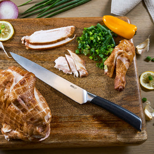 The Zyliss Comfort Pro Carving Knife 20cm rests on a wooden board with sliced smoked chicken, chopped green onions, a yellow pepper, lemon slices, garlic cloves, and green onion stalks.
