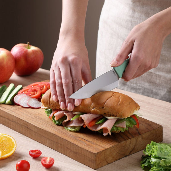 A person uses the Zyliss Classic Utility Knife to slice a sub sandwich filled with lettuce, ham, and tomatoes on a wooden cutting board, with sliced vegetables, an apple, and an orange slice nearby.