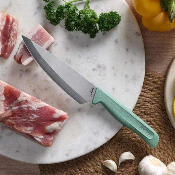 A Zyliss Classic Utility Knife with a green handle rests on a marble cutting board beside raw meat, parsley, garlic, a yellow bell pepper, and a plate, all arranged on a woven placemat.