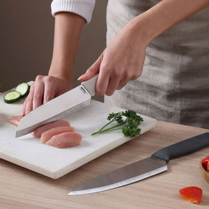 A person slices raw chicken on a white cutting board using a sharp stainless steel chef knife from the Zyliss Classic 2 Piece Chef & Santoku Knife Set. Cucumber slices, parsley, a tomato piece, and the santoku knife from the same Zyliss set are also on the board.