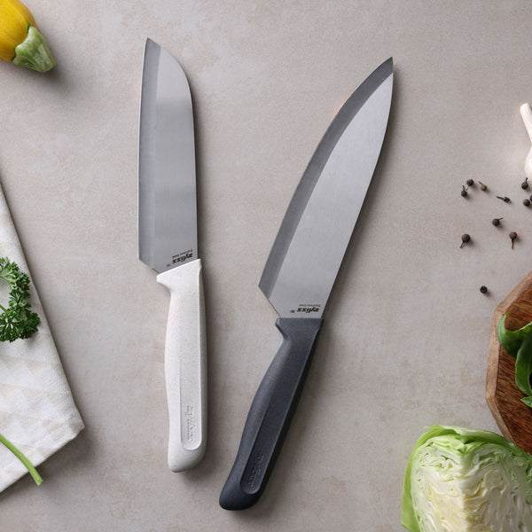 The Zyliss Classic 2 Piece Chef & Santoku Knife Set—featuring one knife with a white handle and the other with a black—rests on a light countertop near cabbage, parsley, yellow squash, scattered peppercorns, and a towel. Each knife is expertly crafted from high-quality stainless steel and includes both a classic chef knife and a versatile santoku blade to meet all your culinary needs.
