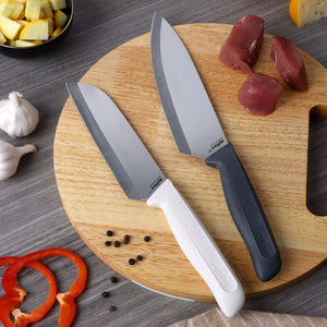A Classic 2 Piece Chef & Santoku Knife Set by Zyliss—one with a white handle and a classic chef knife blade, and the other featuring a black handle and versatile santoku blade—is displayed on a round wooden cutting board. The sharp stainless steel blades are surrounded by raw meat, garlic, cheese, black peppercorns, pepper slices, and freshly chopped vegetables.