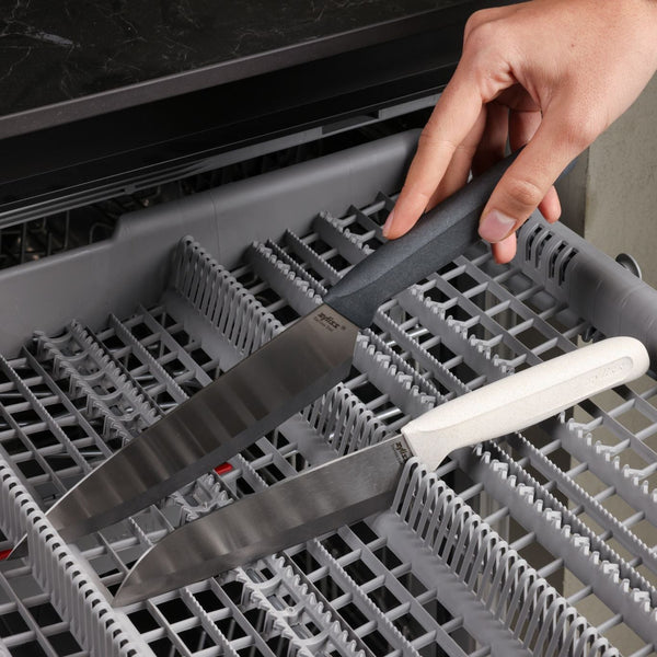 A hand places a Zyliss Classic 2 Piece Chef & Santoku Knife with a black handle into a dishwasher rack, next to another Zyliss knife with a white handle from the same set already inside.