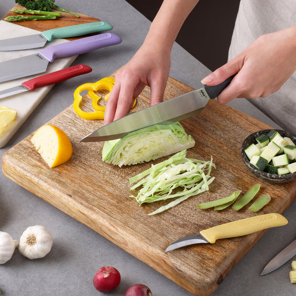 A person slices cabbage on a wooden board surrounded by yellow bell pepper, zucchini, garlic, and radishes. The Zyliss Classic 6-Piece Knife Set—a kitchen essential with stainless steel knives—rests on the counter.