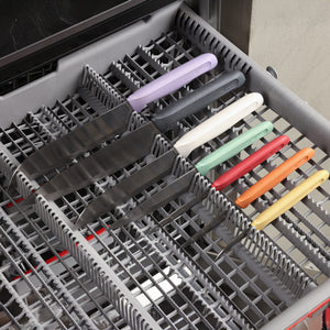 Six vibrant Zyliss Classic knives, each with a different handle color, are neatly arranged blade-down in the top rack of a dishwasher, each in its designated slot.