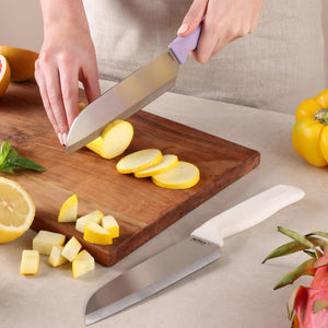 Using the Zyliss Classic 2 Piece Santoku Knife Set, an individual slices yellow squash on a wooden cutting board. The large Santoku knives—each with an ergonomic handle—rest nearby, alongside diced squash, a lemon half, basil, a yellow bell pepper, and another stainless steel knife on a light-colored table.
