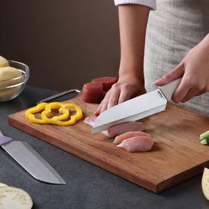 A person slices raw chicken on a wooden cutting board with a stainless steel knife featuring an ergonomic handle from the Zyliss Classic 2 Piece Santoku Knife Set. Slices of yellow bell pepper, raw meat, potatoes, and pieces from the Zyliss Santoku Knife Set are also arranged on the countertop.