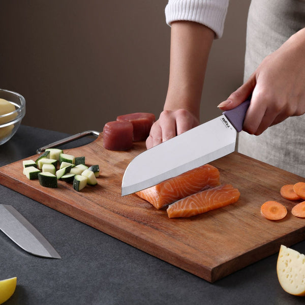 Using the Zyliss Classic 2 Piece Santoku Knife Set, a person slices raw salmon on a wooden cutting board, surrounded by chopped zucchini, carrots, and tuna, with one ergonomic-handled knife in hand while another stainless steel blade from the set rests on the counter.
