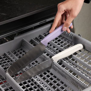 A hand places a large kitchen knife with an ergonomic lavender handle into a dishwasher rack next to a similar knife with a white handle, both from the Zyliss Classic 2 Piece Santoku Knife Set, featuring durable stainless steel blades.