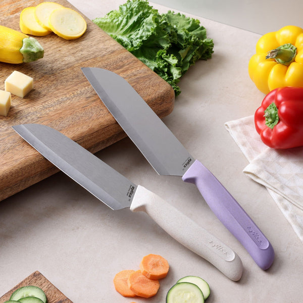 Two kitchen knives from the Zyliss Classic 2 Piece Santoku Knife Set, featuring white and purple ergonomic handles, rest on a counter near sliced vegetables, a cutting board, lettuce, yellow squash, and red and yellow bell peppers. This vibrant scene suggests meal preparation made easy with the durable stainless steel blades of the Zyliss Classic Santoku Knife Set.