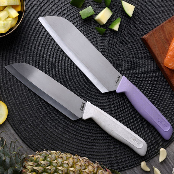 Two kitchen knives from the Zyliss Classic 2 Piece Santoku Knife Set—featuring silver stainless steel blades and ergonomic pastel handles in white and lavender—are displayed on a black textured mat alongside cut vegetables, fresh pineapple, and a wooden board topped with salmon. Designed by Zyliss for comfort and precision, the sharp stainless steel blades make every culinary task effortless.