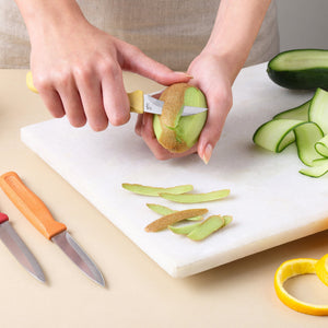 A person uses a small knife from the Zyliss Classic 3 Piece Peeling & Paring Knife Set to peel a kiwi on a white cutting board, with additional prep knives and cucumber slices nearby.
