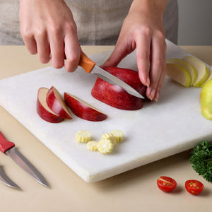 A person slices a red apple on a white cutting board with a stainless steel paring knife from the Zyliss Classic 3 Piece Peeling & Paring Knife Set. Nearby sit a partially sliced apple, corn pieces, halved tomatoes, and the rest of their Zyliss kitchen prep knives.